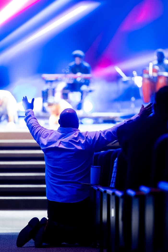 A man kneels with arms raised during a live worship concert with vibrant lighting.